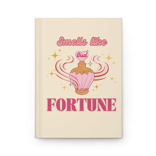 light pastel yellow hardcover journal with an illustration of a pink vintage oud perfume bottle and a retro aesthetic baby pink text "smells like fortune"